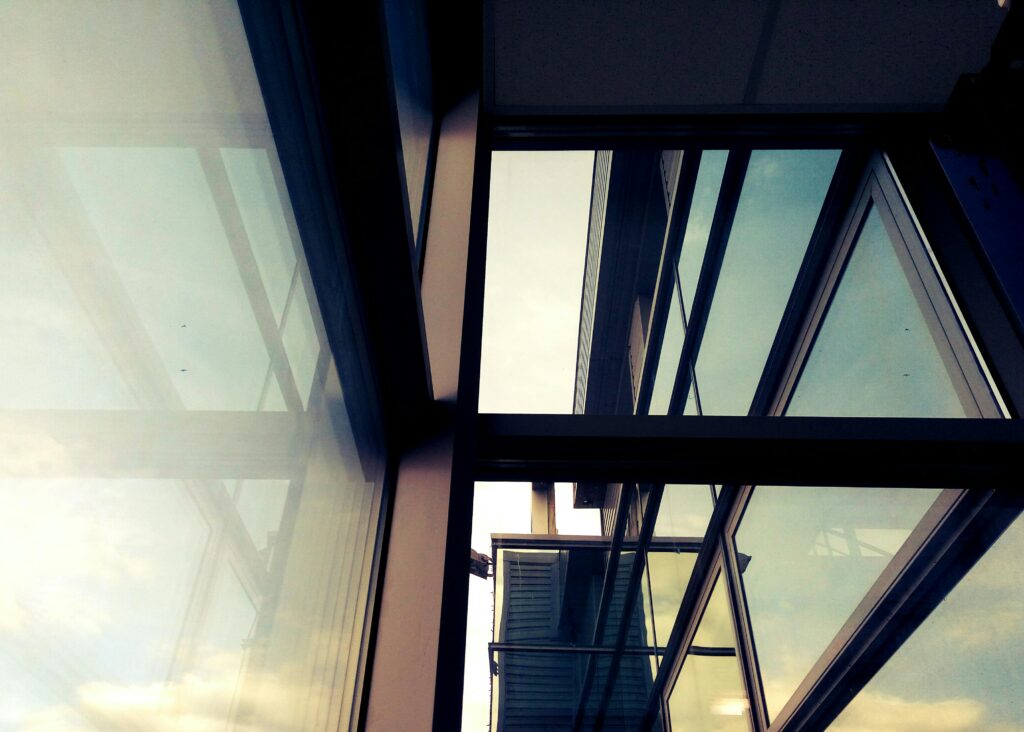 A modern building's glass and steel structure with reflections, captured indoors during the day.